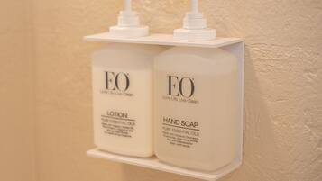 To flaske EO lotion og håndsæbe står på en hvid reol foran en væg med struktur.