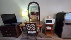 A compact hotel room with a flat-screen TV, a wooden desk, a chair, a microwave, a refrigerator, a bedside lamp, and a mirror. - Gulf Way Inn (Clearwater)