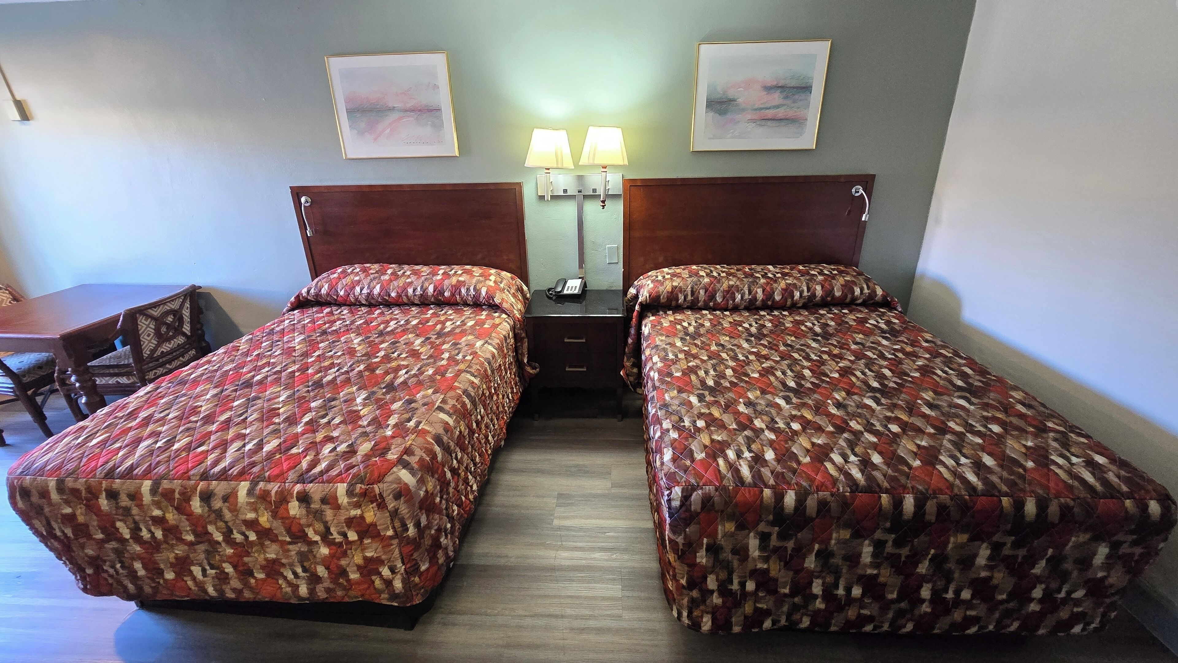 Une chambre d’hôtel avec deux lits simples, une tête de lit en bois, une table de chevet avec une lampe et un bureau avec une chaise.