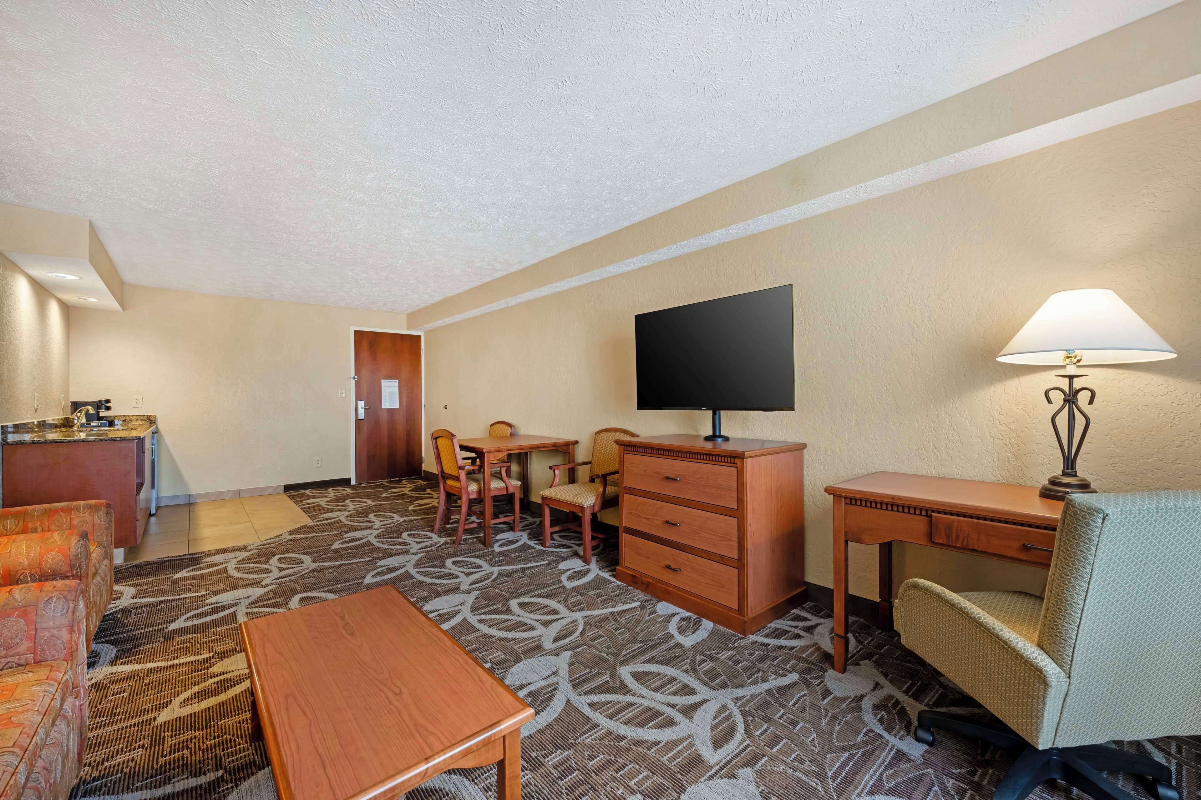 A hotel room with a kitchenette, a flat-screen TV, a desk with a lamp, a chair, and a sofa.