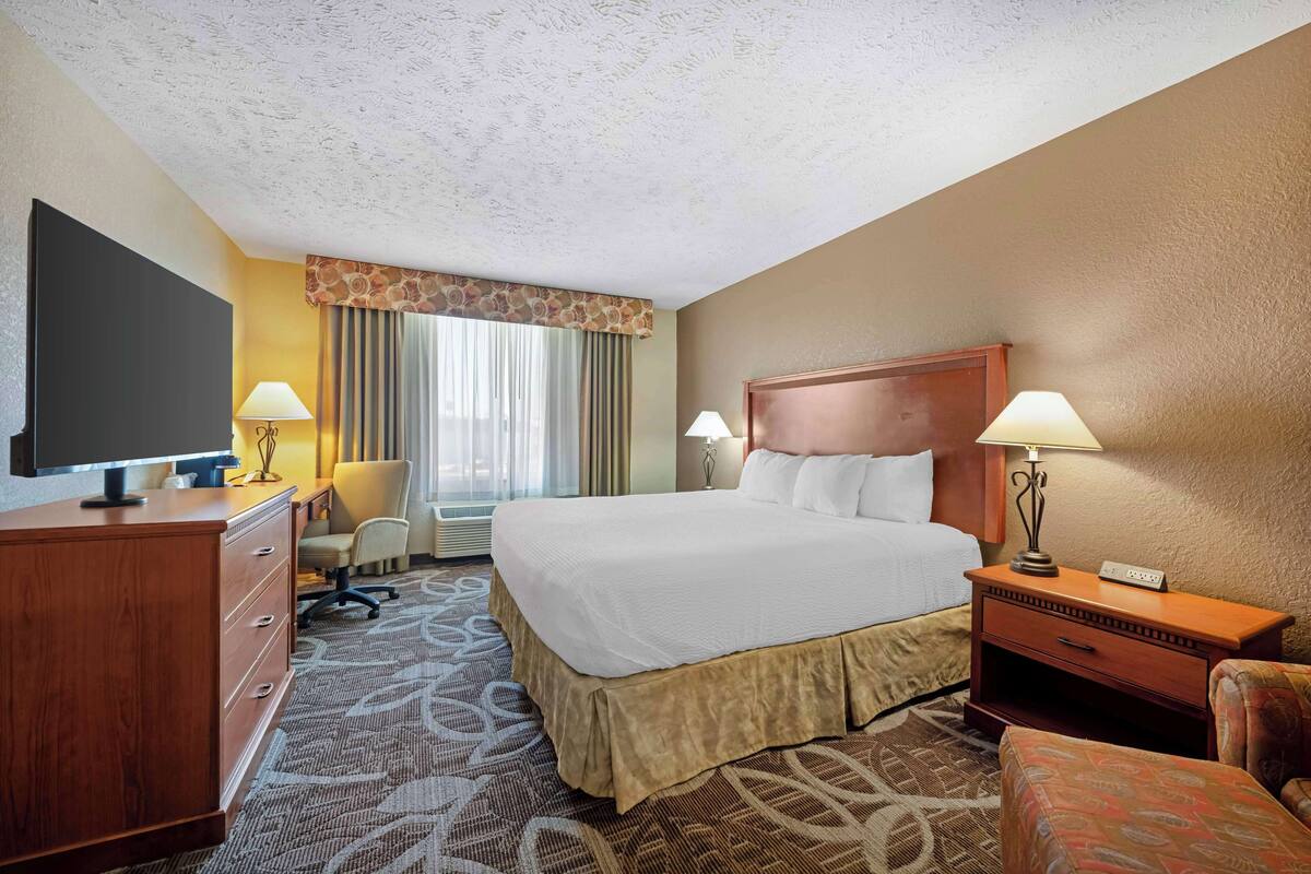 A hotel room with a large bed, a desk with a lamp, a television, and a patterned carpet.