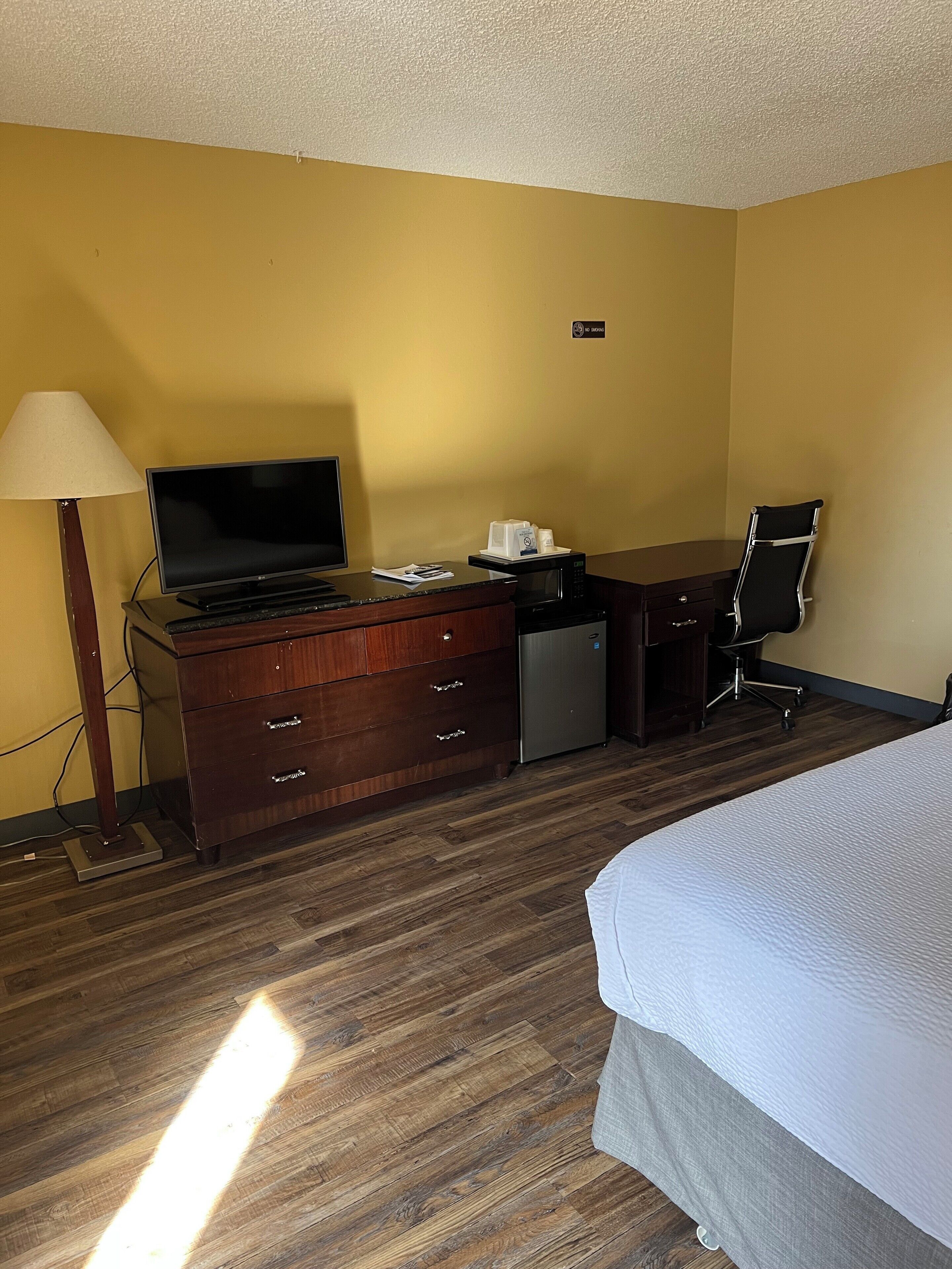 A hotel room with a bed, a desk with a computer, a lamp, a microwave, and a chair.