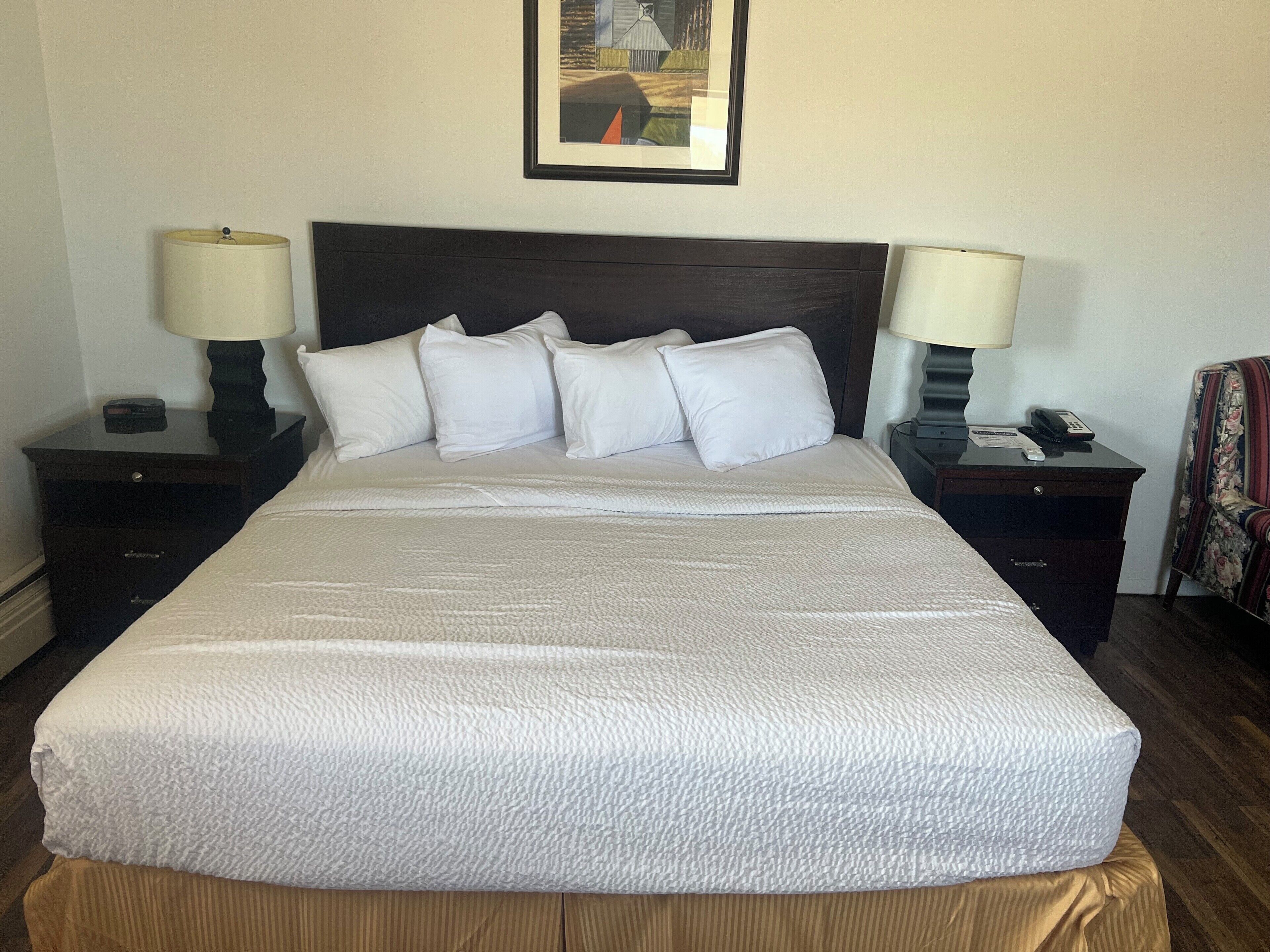 A neatly made bed with a white comforter and pillows, flanked by two bedside tables with lamps, a framed picture above the bed, and a chair with a floral pattern to the right.