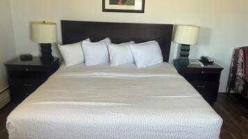 A neatly made bed with a white comforter and pillows, flanked by two bedside tables with lamps, a framed picture above the bed, and a chair with a floral pattern to the right.