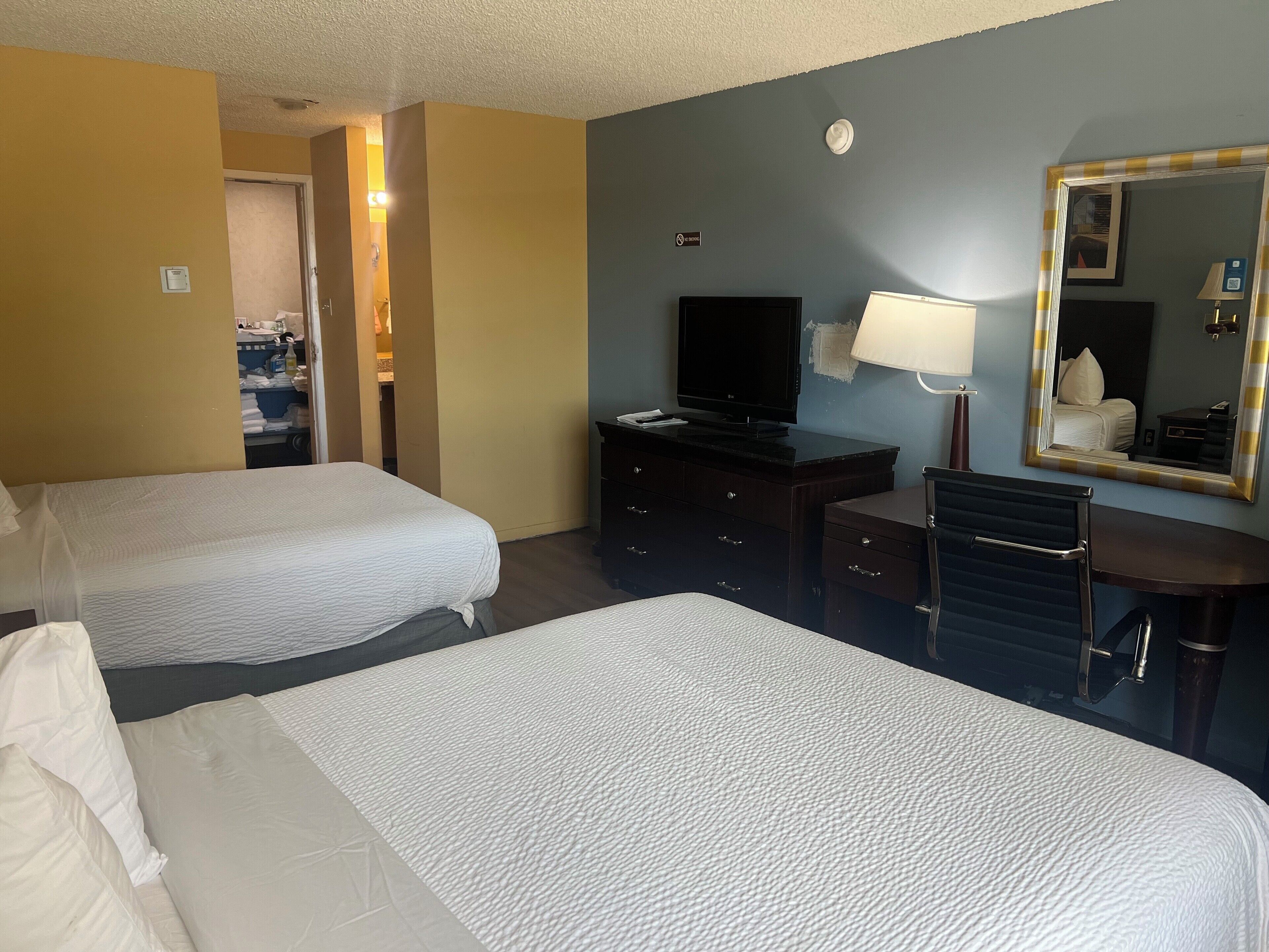 A hotel room with two beds, a desk, a television, a mirror, and a bathroom visible through an open door.