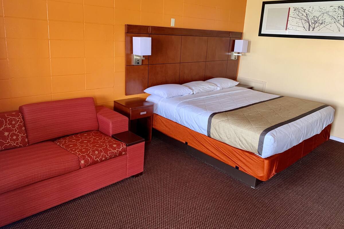 A hotel room with a bed, a red sofa, a nightstand, and a wall-mounted lamp.