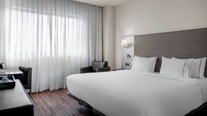 Habitación de hotel con una cama grande, un escritorio, una silla y una ventana con cortinas traslúcidas.