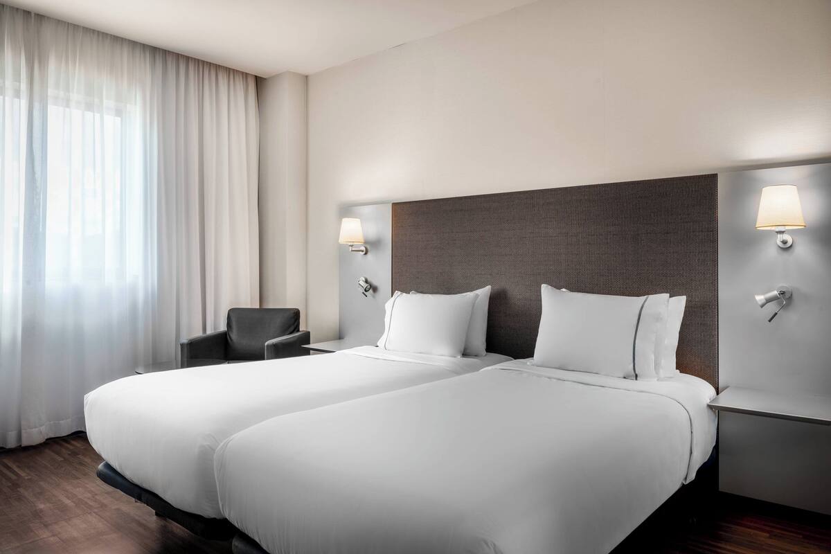 Een hotelkamer met twee bedden, een stoel, een nachtkastje en een groot raam met gordijnen.