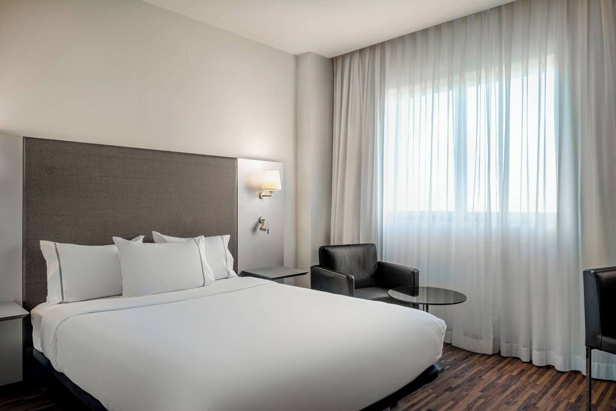 Een moderne hotelkamer met een groot bed, een hoofdeinde, een nachtkastje met een lamp, een klein rond tafeltje, een stoel en een groot raam met dunne gordijnen.