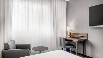 A hotel room with a bed, a desk with a chair, a small round table, and a TV mounted on the wall.