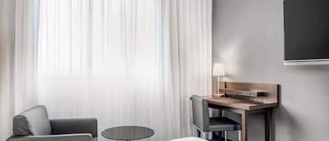 A hotel room with a bed, a desk with a chair, a small round table, and a TV mounted on the wall.