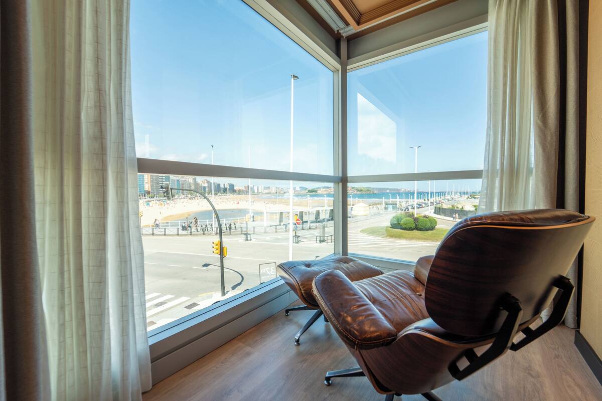 Una habitación con un amplio ventanal que da a un paisaje urbano, un sillón de cuero y suelo de madera.