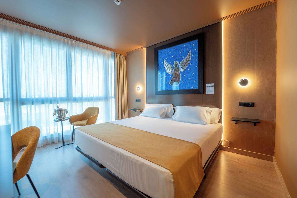 Habitación de hotel con una cama grande, un escritorio con una silla, una mesita con una lámpara y un ventanal con cortinas.