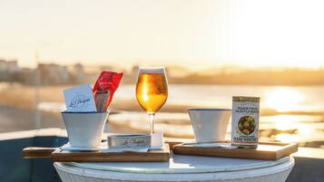 A table with a glass of beer, a packet of snacks, a can of olives, and a notebook on a seaside terrace.