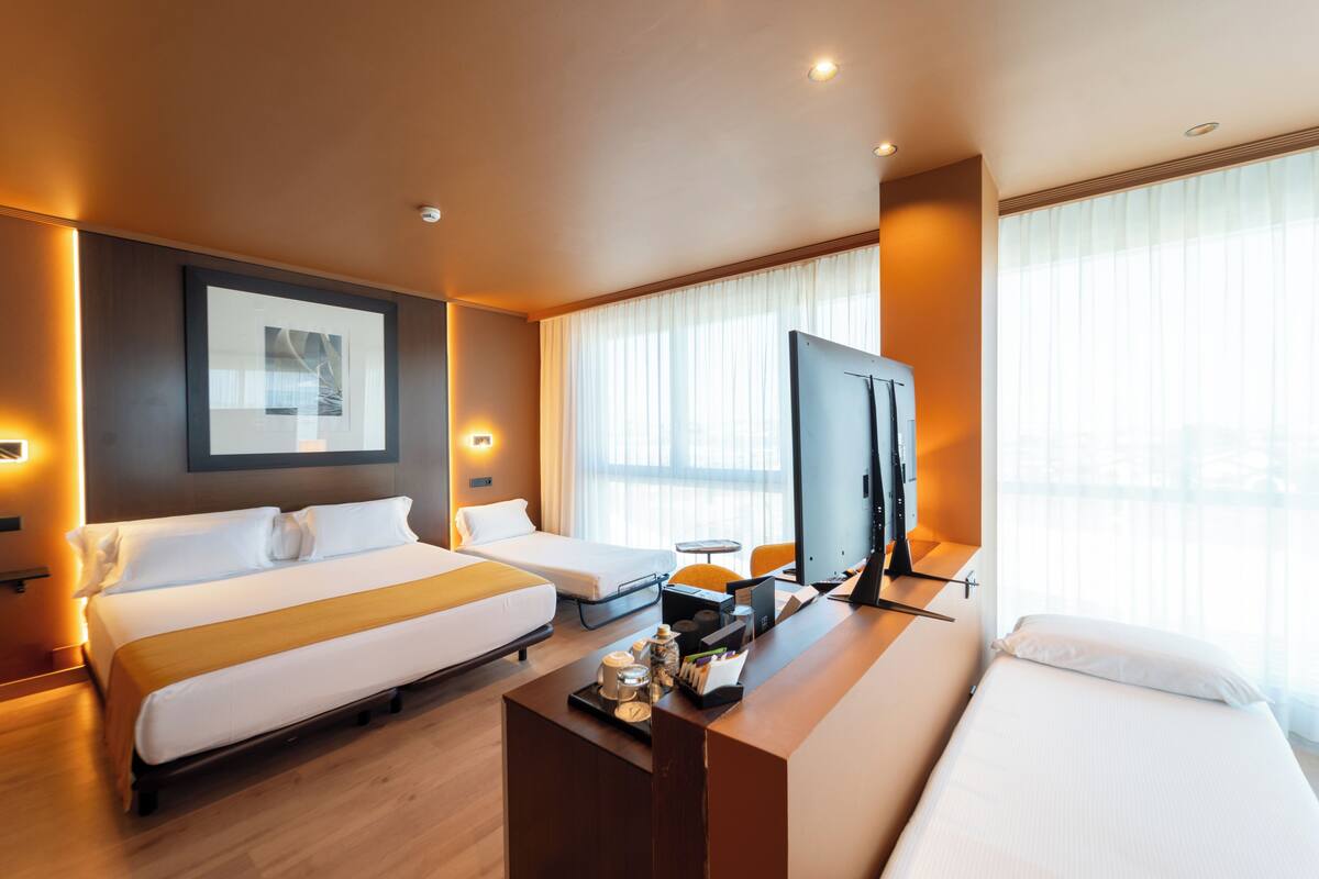 Habitación de hotel con dos camas, un escritorio con televisor y un ventanal con cortinas.