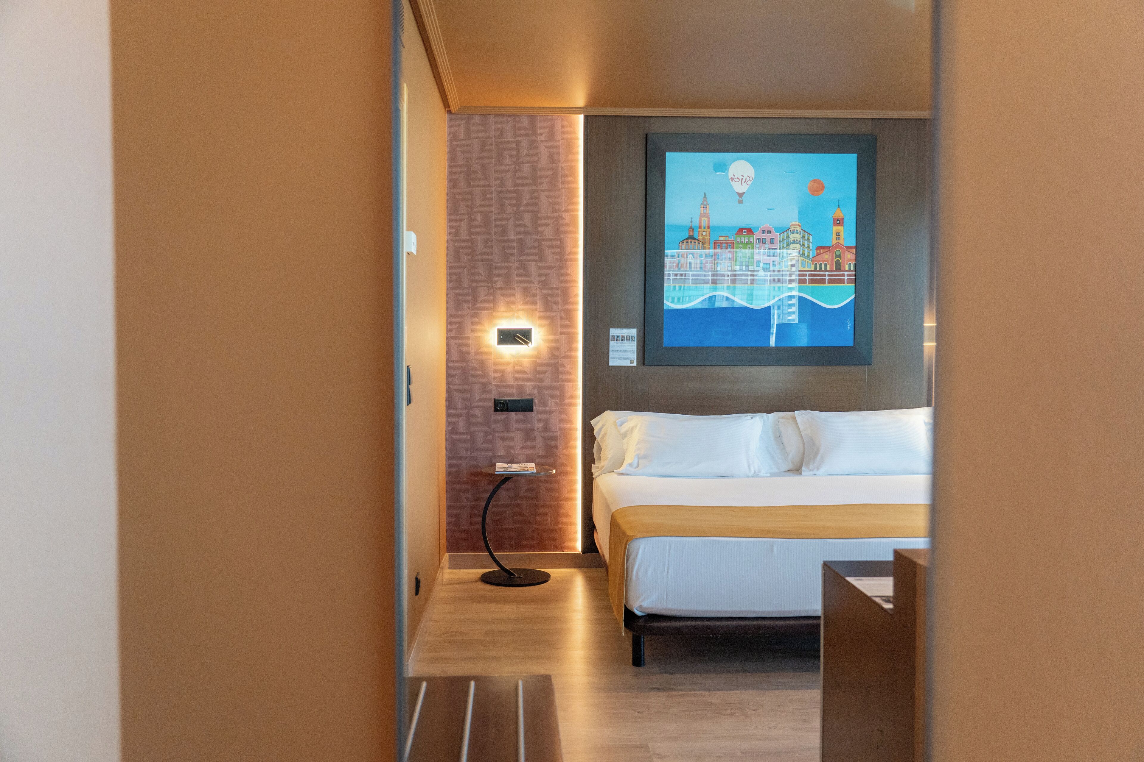 A hotel room with a bed, a nightstand, a wall-mounted TV, and a framed artwork depicting a cityscape.