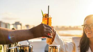 A person holding a cocktail glass with a straw and garnish, with a cityscape in the background.