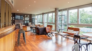 A spacious living area with a wooden floor, a bar area with stools, and a large window offering a view of greenery outside. - Abba Playa Gijon Hotel (Gijon)