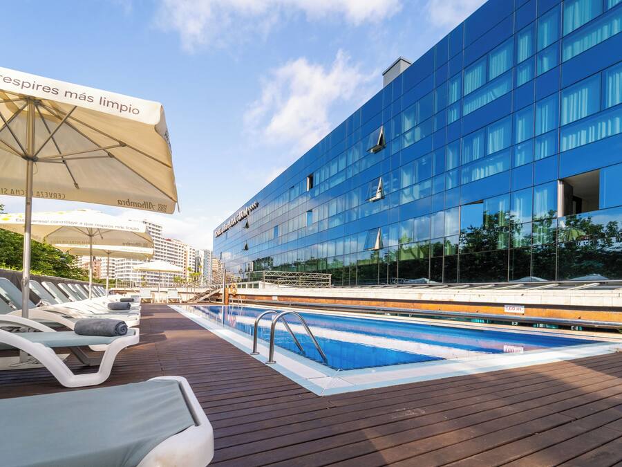 A swimming pool area with lounge chairs and umbrellas, a modern building with reflective glass windows, and a clear sky.
