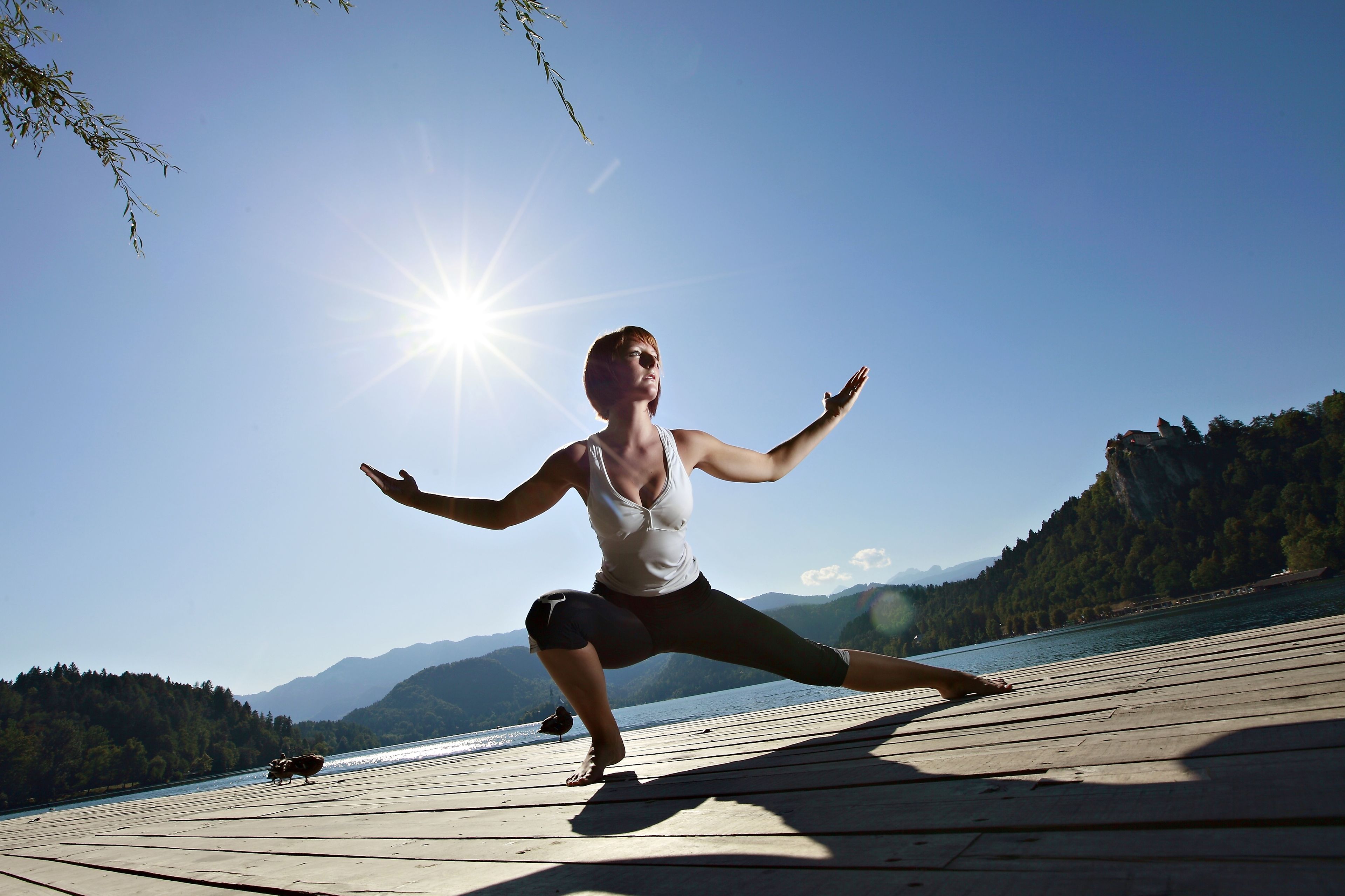 Una persona realiza una postura de yoga sobre una terraza de madera junto a un lago, con montañas al fondo.