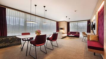 Una habitación de hotel moderna con zona de comedor, un conjunto de sofás y un amplio ventanal con cortinas.