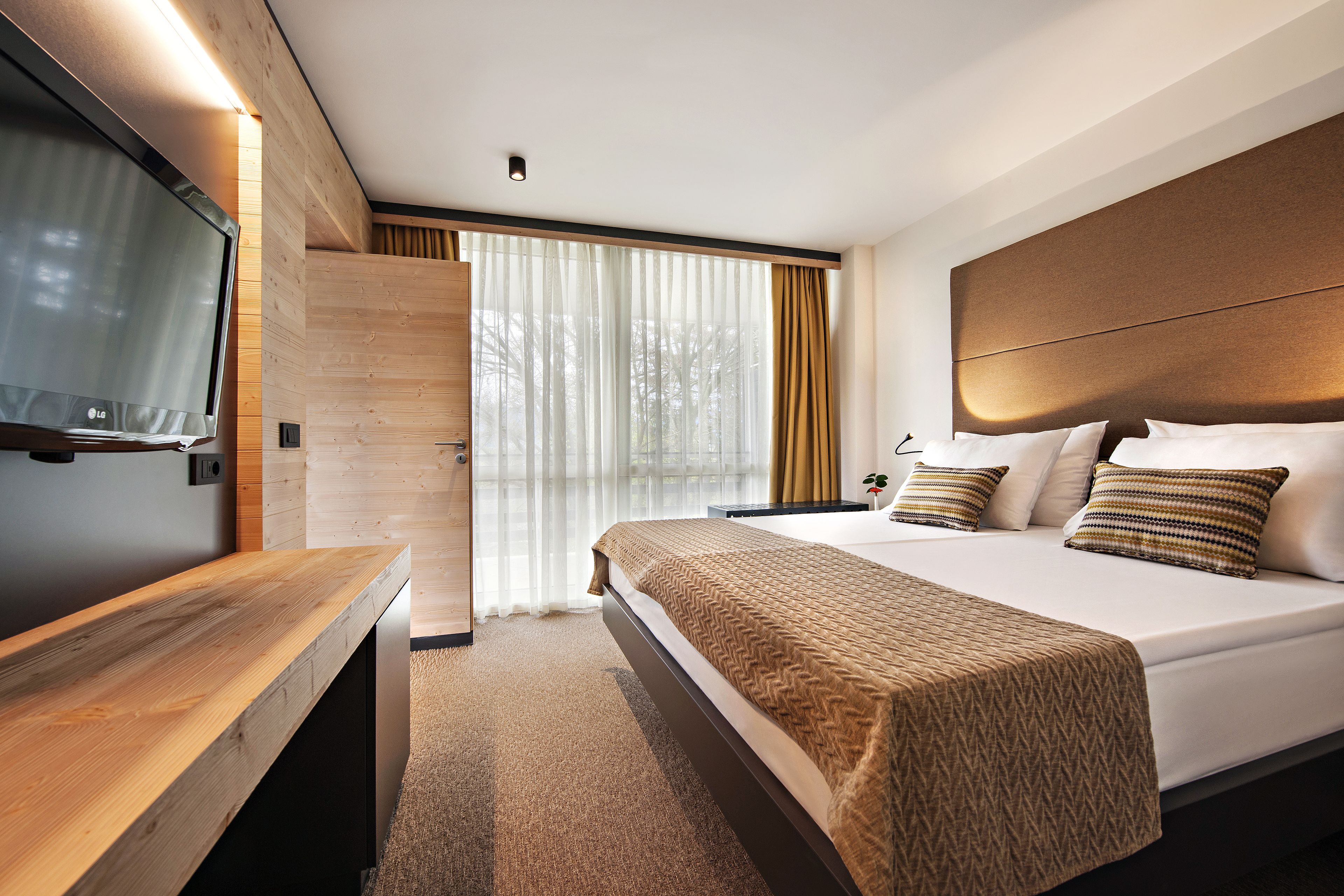 A modern hotel room with a large bed, a wooden desk, a flat-screen TV, and a large window with curtains.