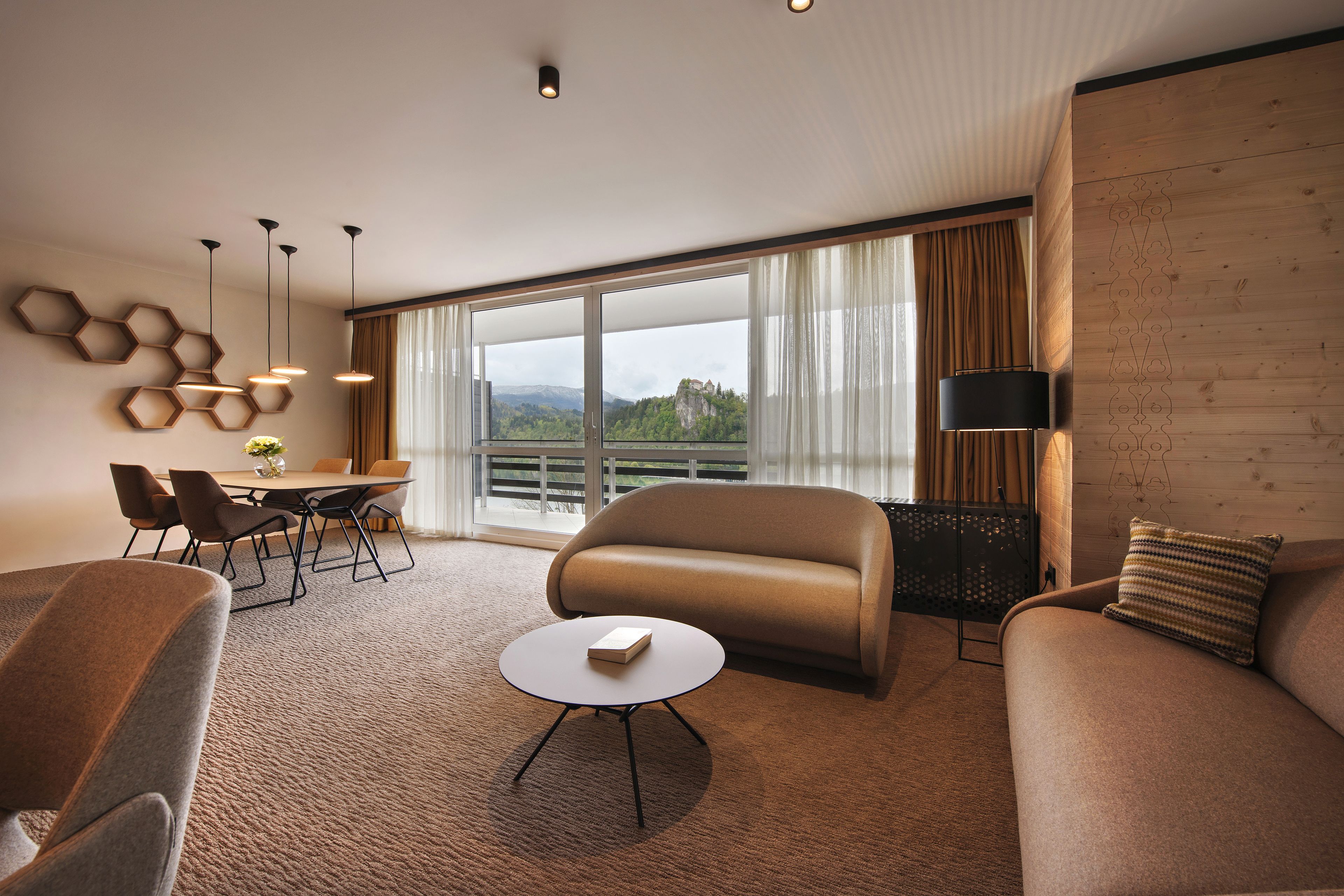 Ein modernes Hotelzimmer mit einer Couch, einem Sessel, einem Esstisch und Stühlen. Ein großes Fenster bietet Blick auf eine gebirgige Landschaft.