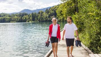 Zwei Personen gehen über ein Holzsteg auf einem See, umgeben von Grünflächen und Bergen.