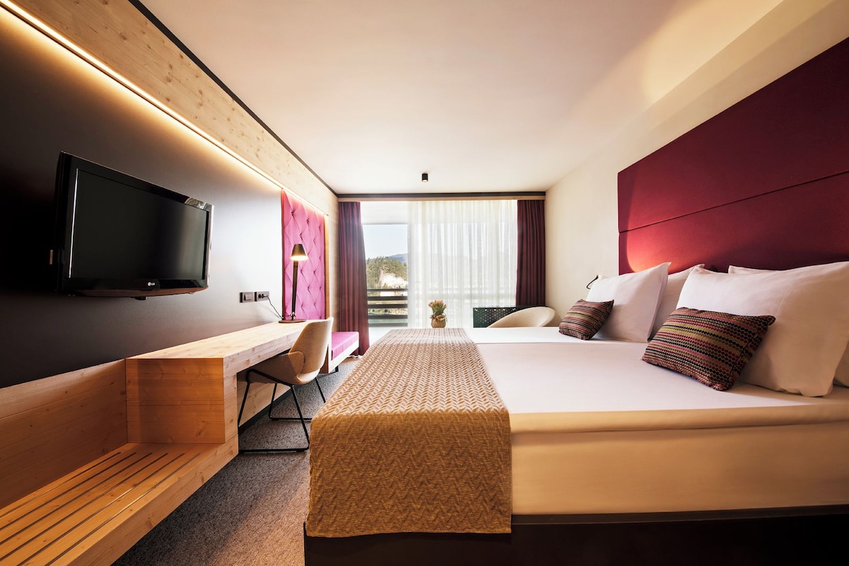A modern hotel room with a large bed, a desk, a chair, and a TV.