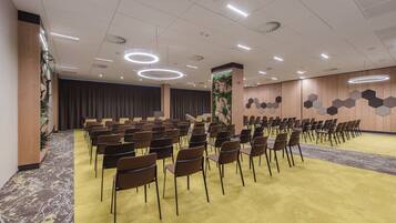 Un salón de conferencias con filas de sillas, piso alfombrado y un escenario con cortinas.