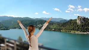 Una mujer con los brazos levantados, mirando un lago con montañas al fondo.