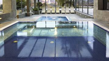 An indoor swimming pool with clear water, surrounded by lounge chairs and a view of a cityscape through large windows.