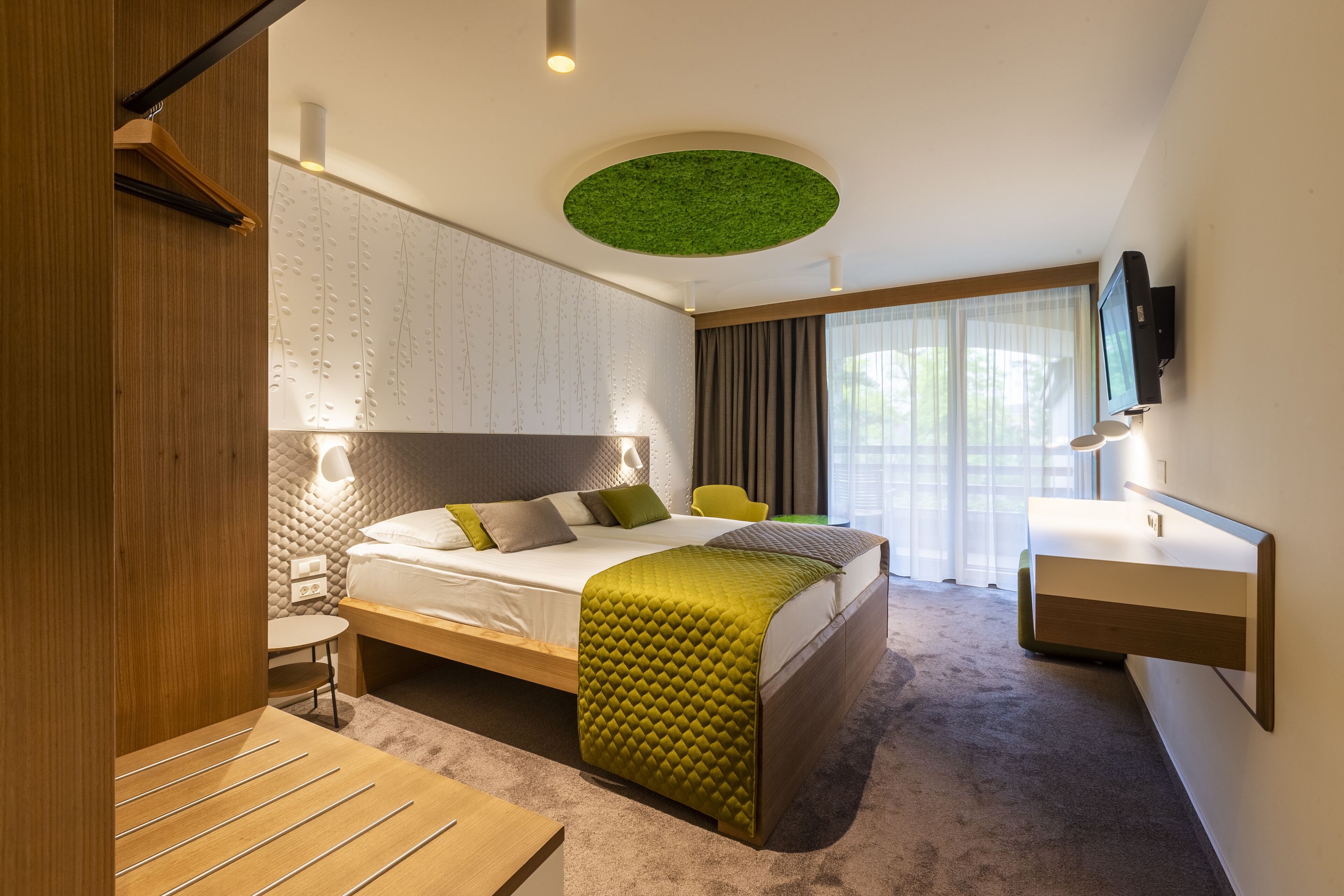 Habitación de hotel moderna con una cama grande, una mesita de noche, un escritorio, una silla, un televisor y un elemento decorativo en el techo verde.