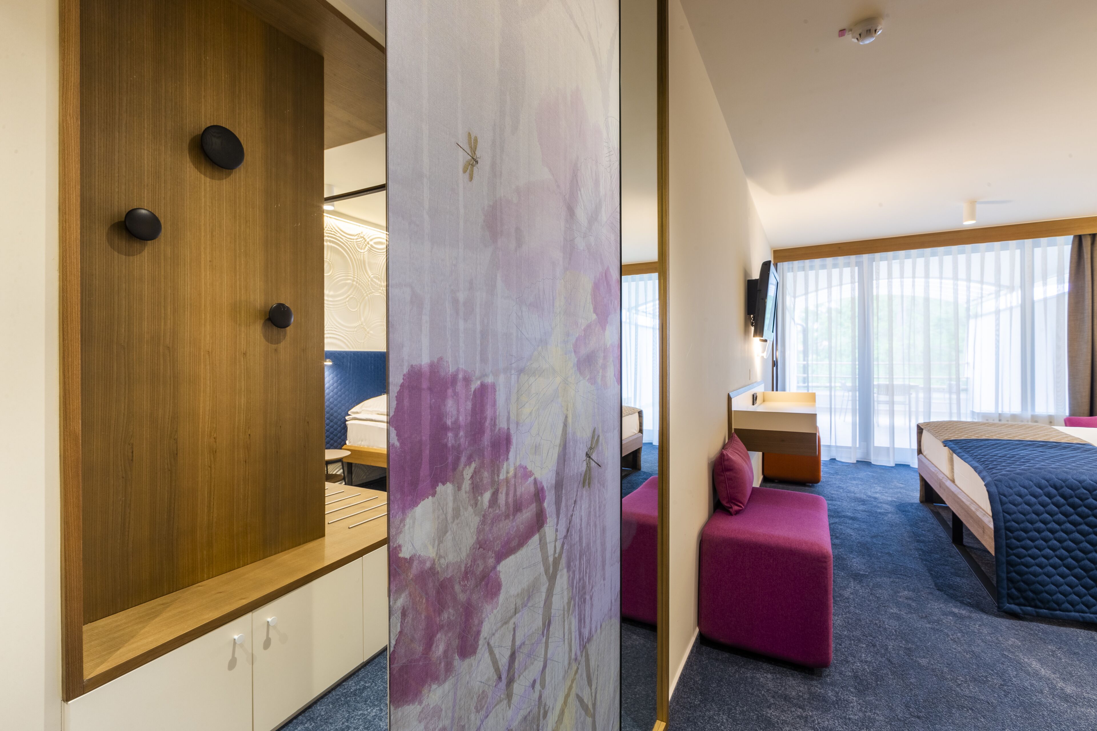 A modern hotel room with a wooden wardrobe, a large mirror, a pink ottoman, a bed with a quilted cover, and a large window with sheer curtains.