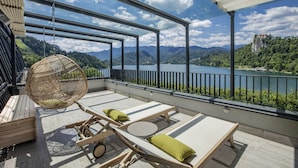 Ein Balkon mit Liegestühlen, einem Korbsessel und Blick auf einen See und Berge.