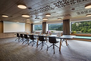 A conference room with a large table, chairs, a projector screen, and large windows with a view of greenery.