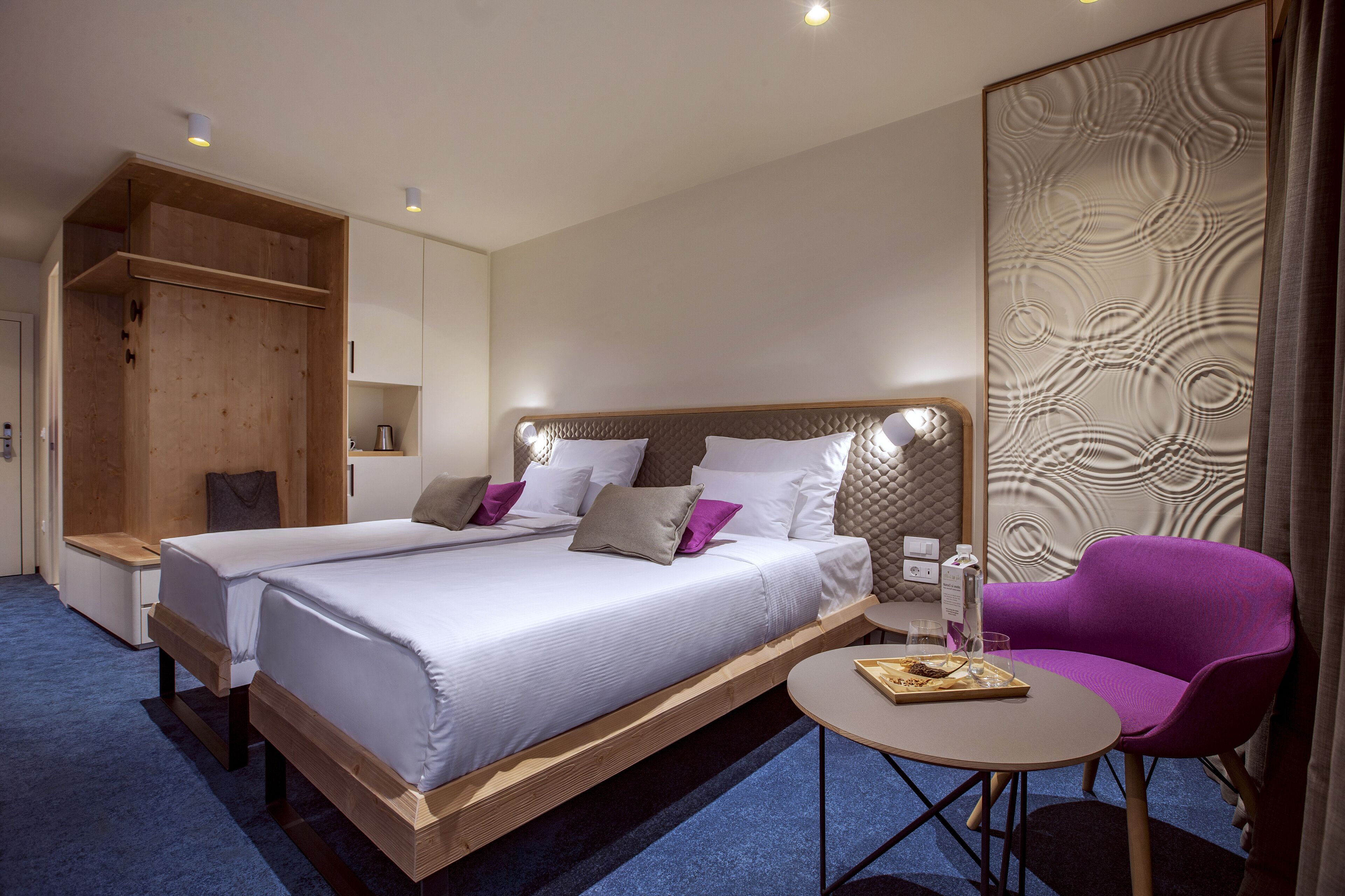Ein Hotelzimmer mit einem großen Bett, einem kleinen Tisch, einem violetten Stuhl und einem Holzschrank.