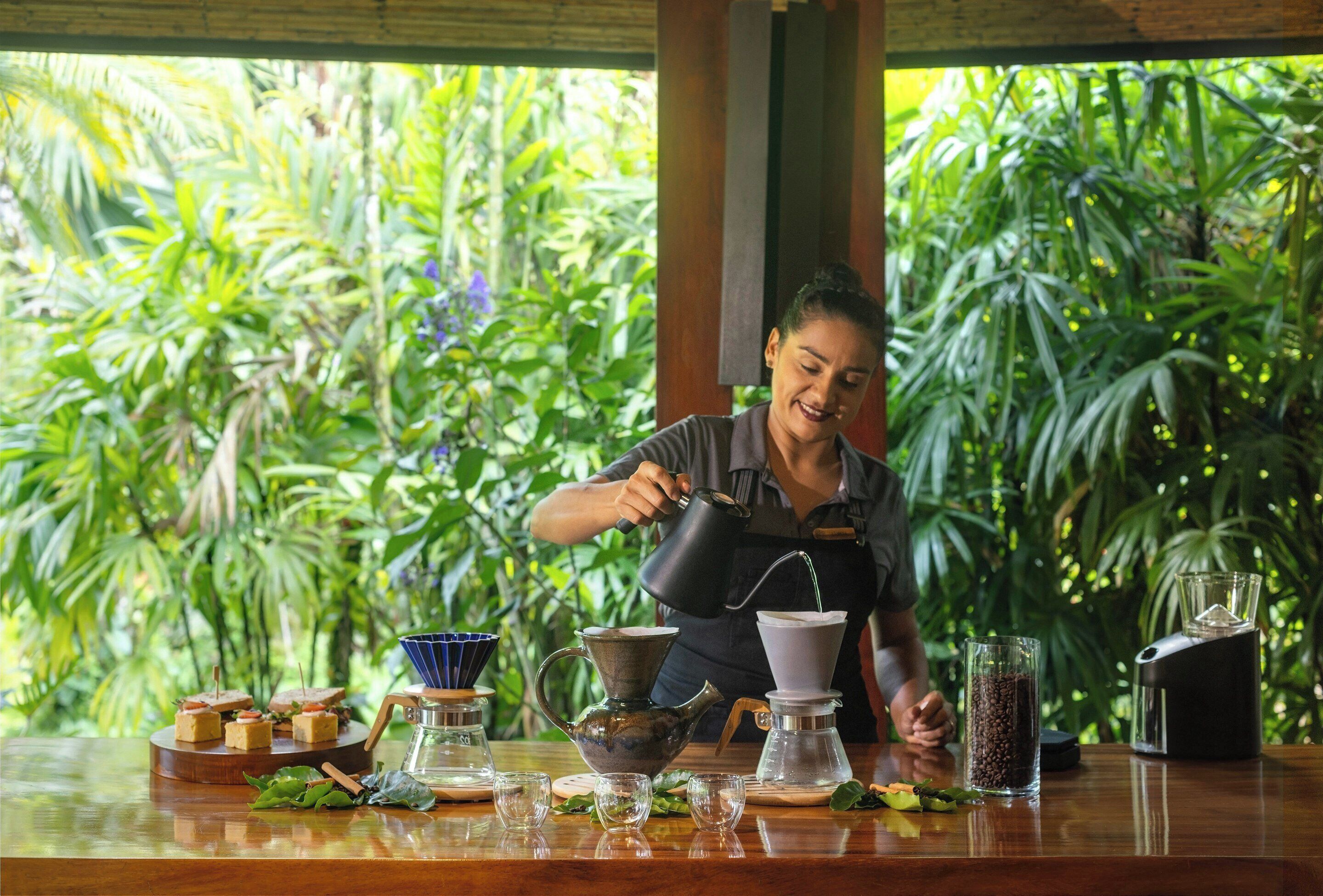 Eine Person gießt Kaffee in ein Glas, mit einer Kaffeemaschine und verschiedenen Kaffeeutensilien auf einem Tisch.
