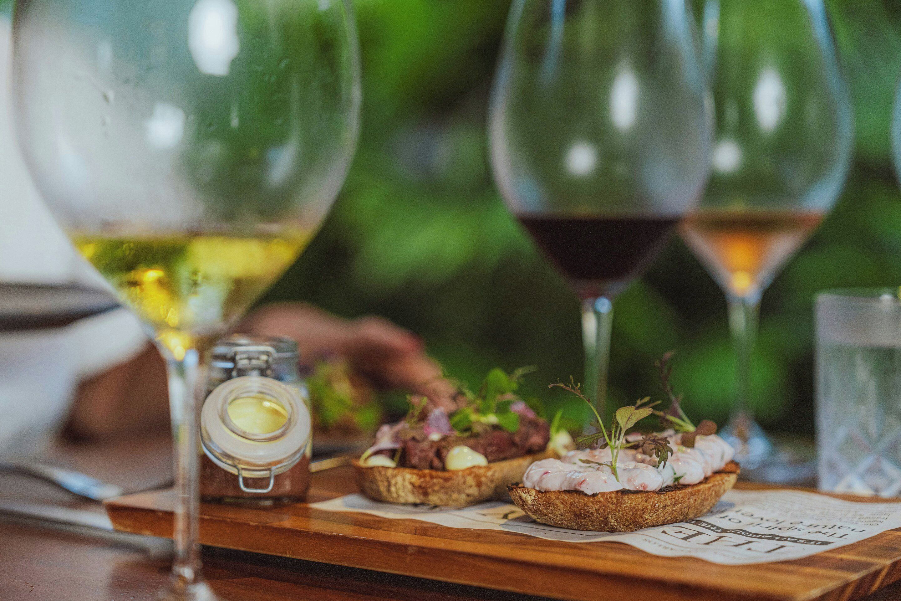 Una mesa de madera con dos copas de vino, un frasco pequeño y dos rebanadas de pan con condimentos.