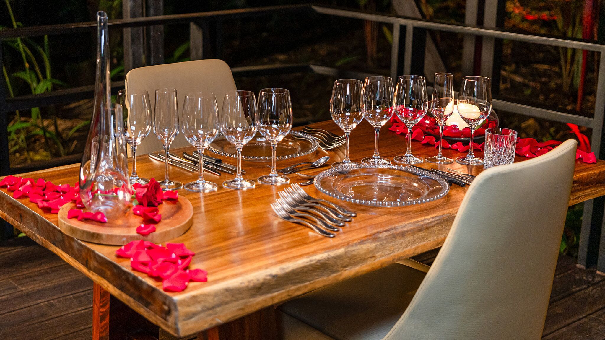 Una mesa de comedor preparada con copas de vino, platos y cubiertos, con pétalos de rosa roja esparcidos sobre la mesa.