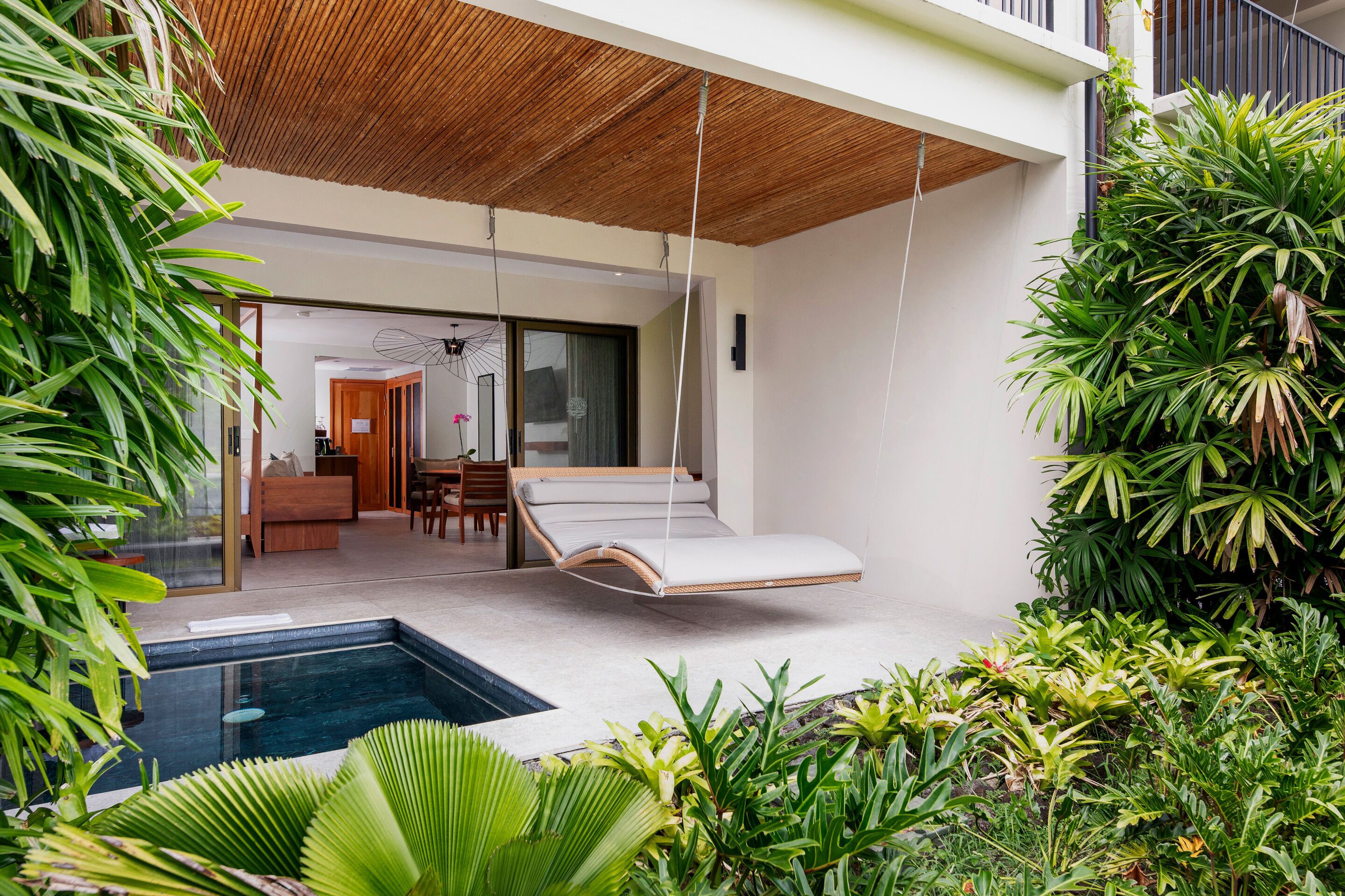 A modern outdoor living area with a hanging lounge chair, a pool, and lush greenery.