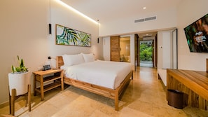 A modern hotel room with a large bed, a bedside table with a lamp, a TV, and a potted plant.