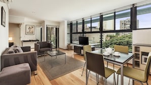 A modern living room with a sofa, chairs, a coffee table, and a TV. There is a large window with a view of buildings and greenery outside.