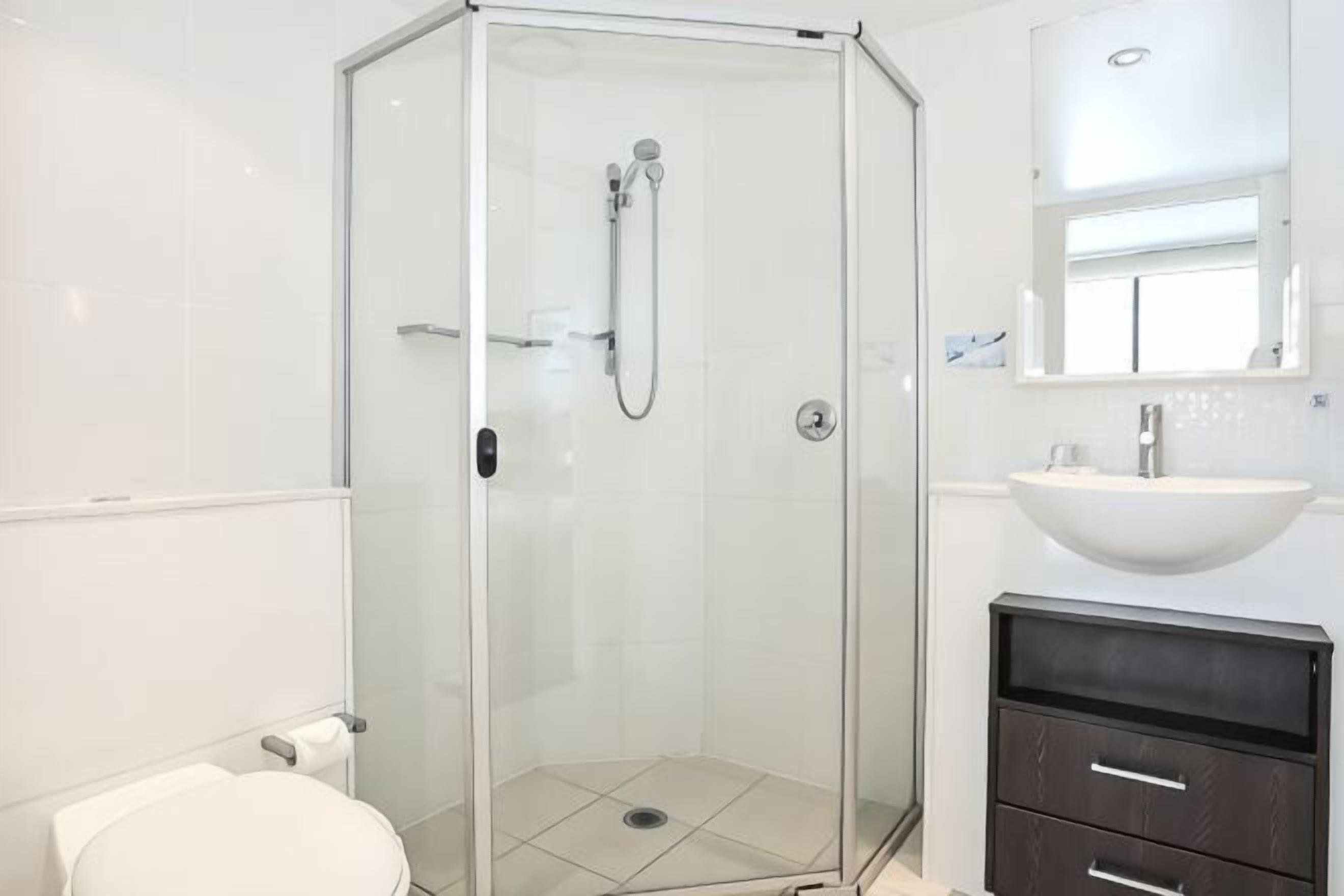 Un baño con ducha de vidrio, lavamanos blanco y tocador oscuro.