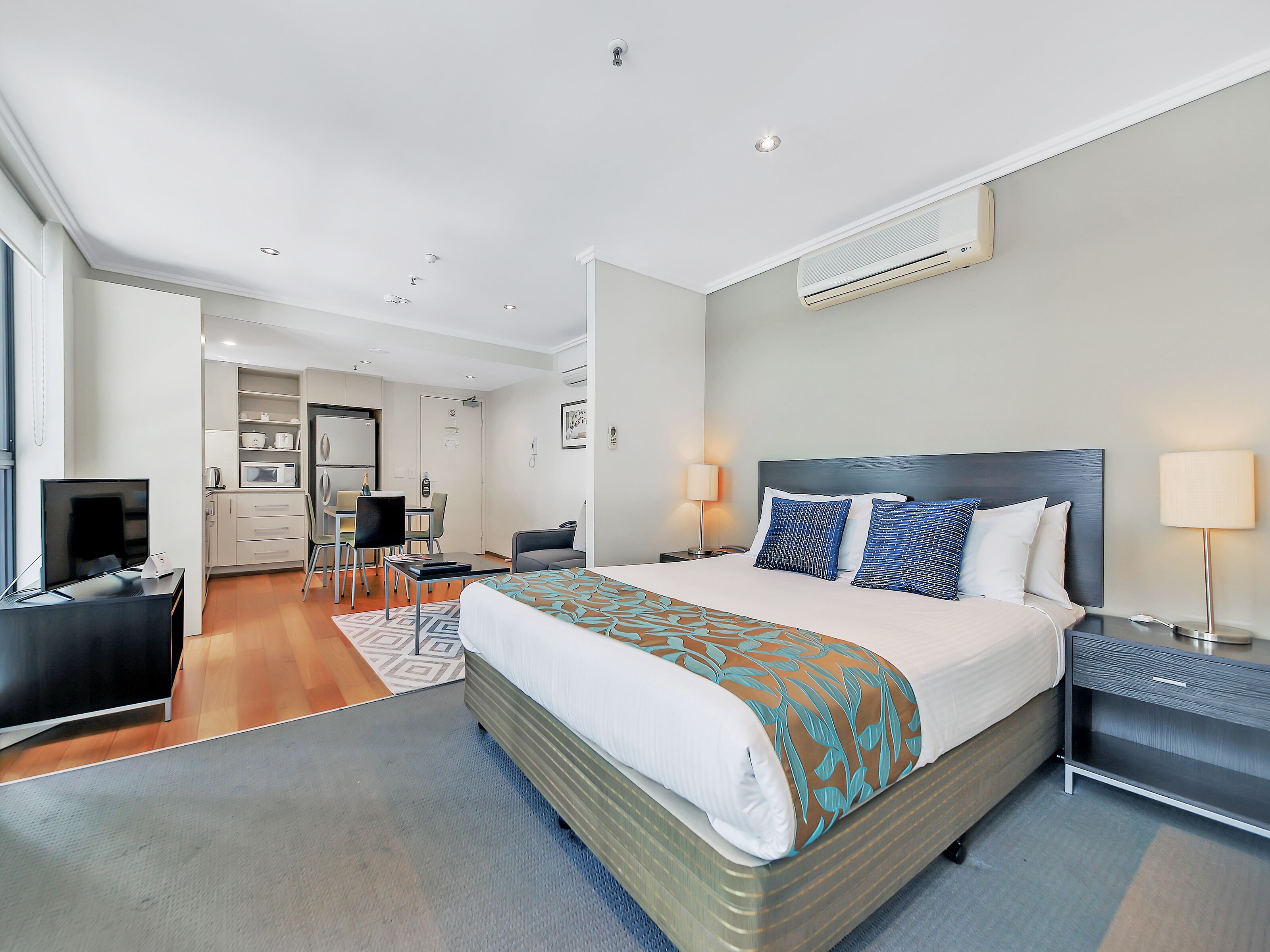 A modern hotel room with a large bed, bedside tables, a desk, and a kitchenette.