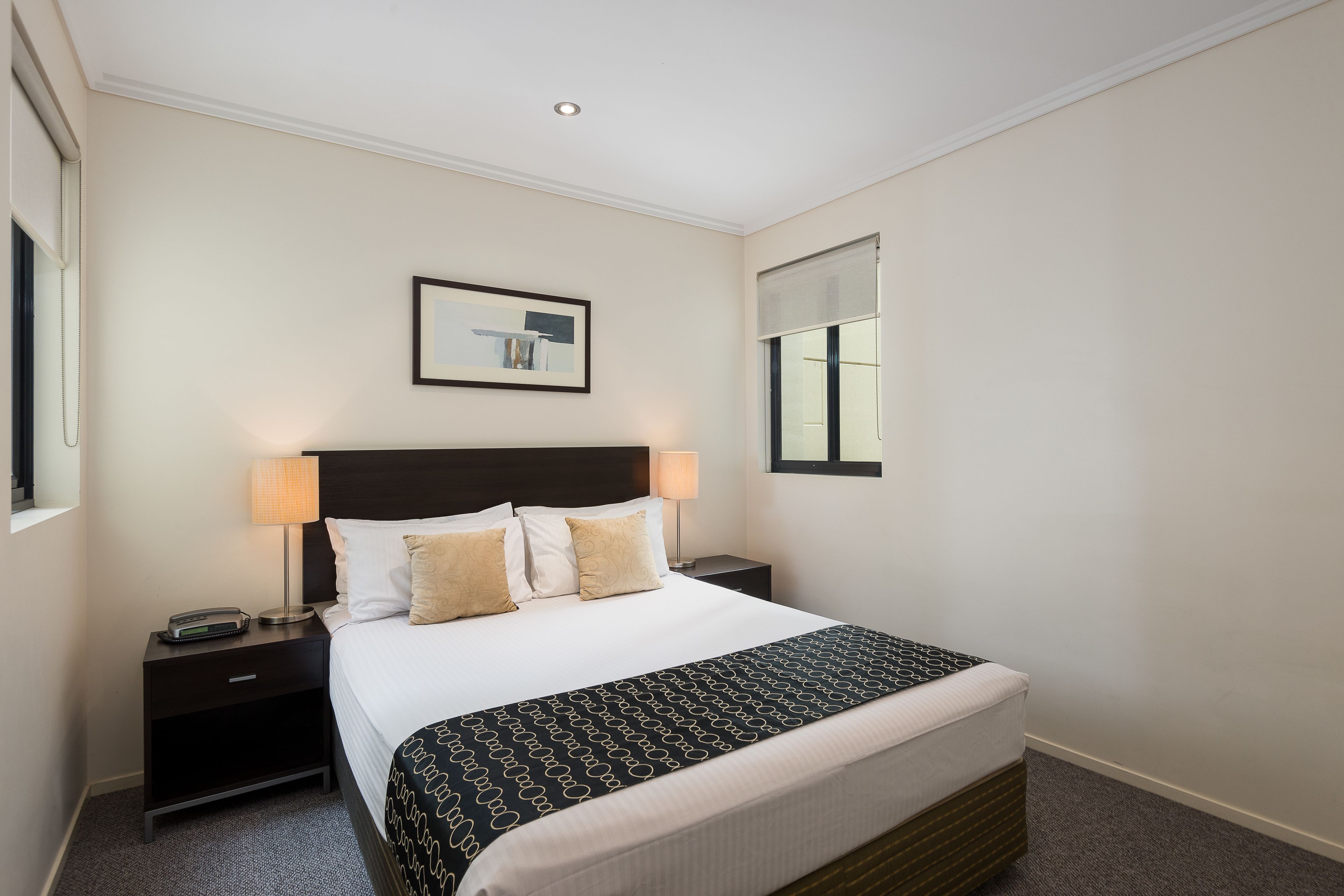 Een hotelkamer met een bed, nachtkastjes, een lamp, een ingelijst schilderij aan de muur en een raam met jaloezieën.