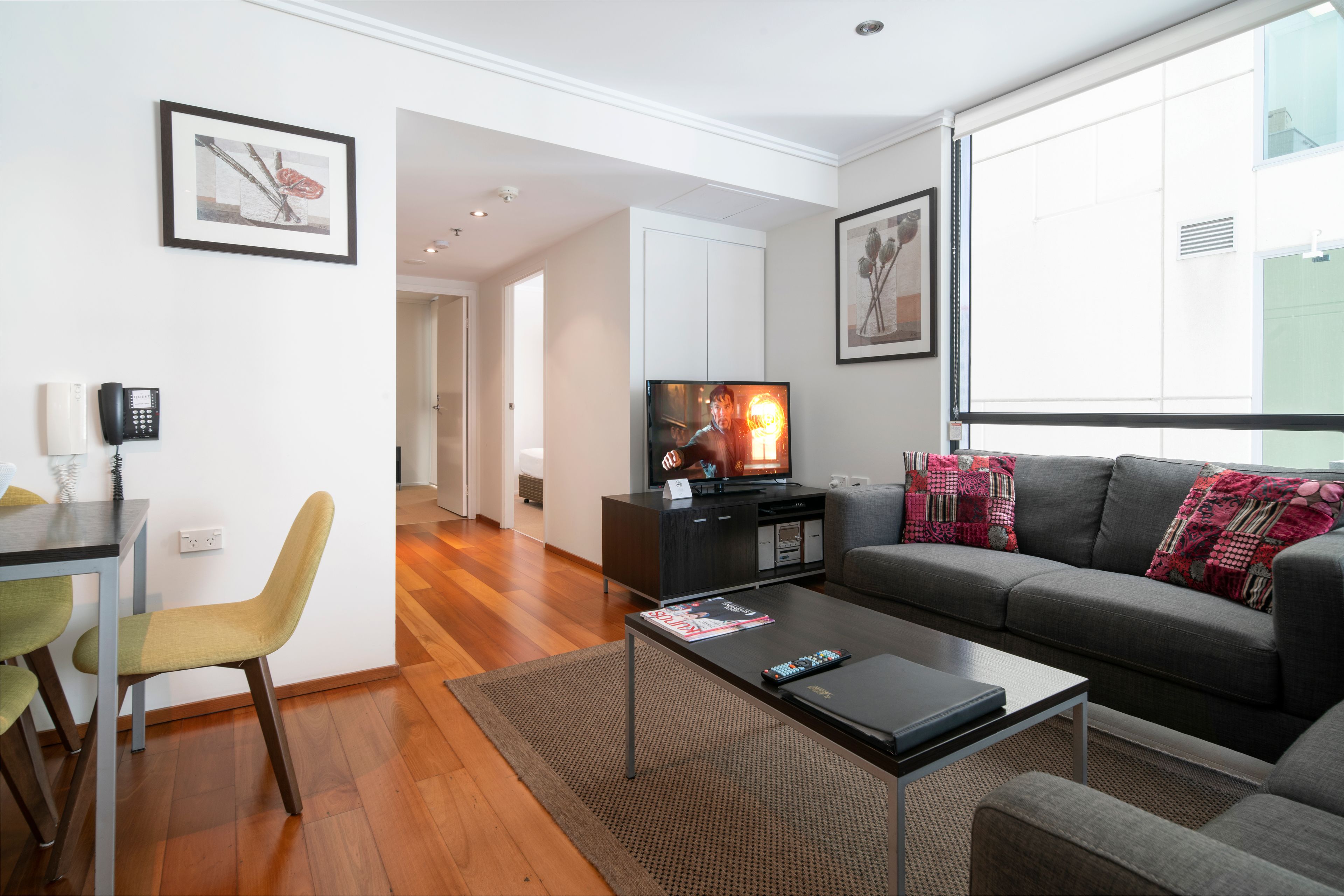 A modern living room with a grey sofa, a coffee table, and a television. There is a dining area with a table and chairs, and a kitchen area with cabinets.