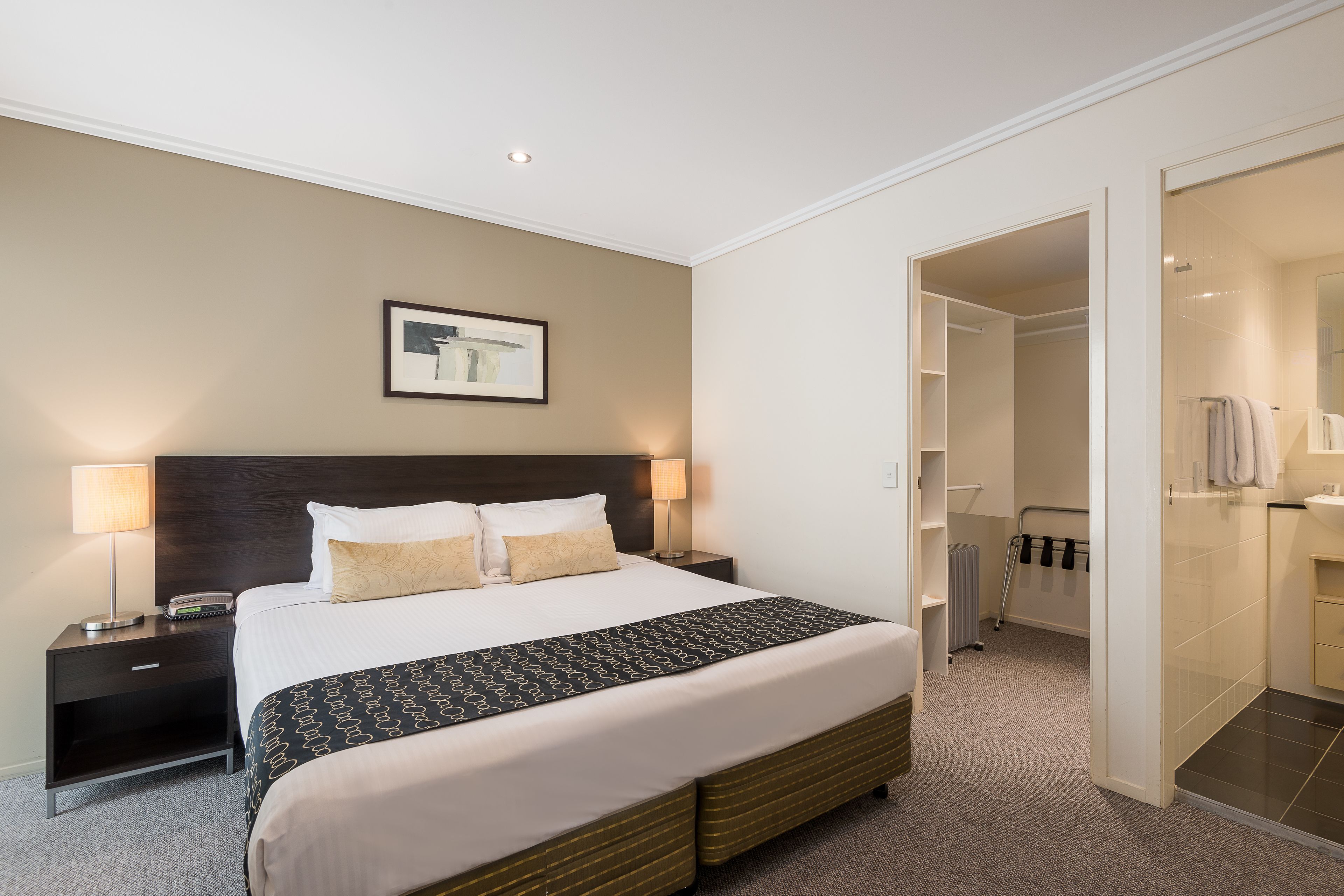 Habitación de hotel con una cama grande, una mesita de noche con lámpara, un cuadro en la pared y un vestidor.