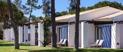 A row of modern villas with white walls and blue shutters, each with a covered patio and two lounge chairs.