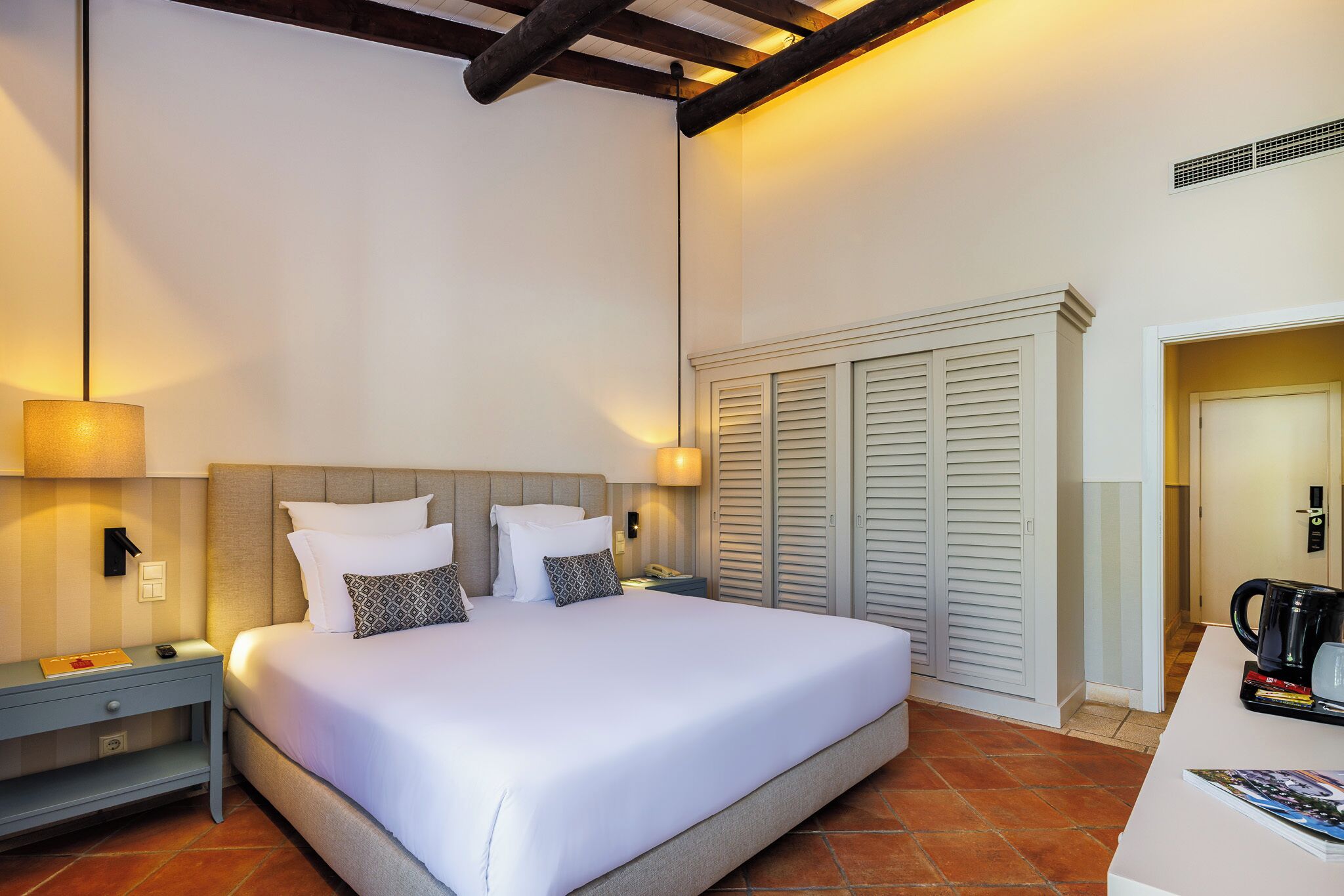 Un lit bien fait, avec du linge de lit blanc et des oreillers, une table de chevet avec une lampe, une commode et une armoire à portes à persiennes.